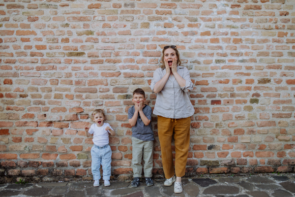 A young mother with her little children standing by brick wall and making funny faces in street summer.