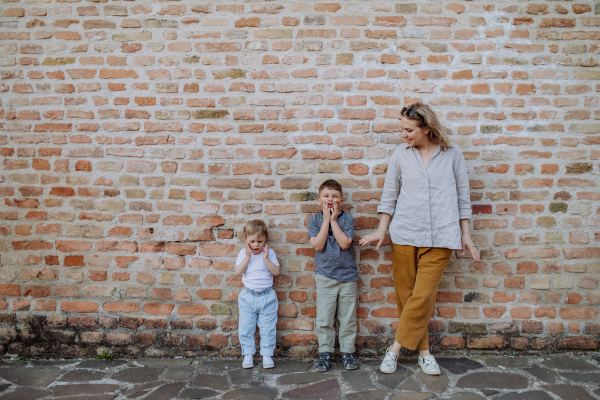 A young mother with her little children standing by brick wall and making funny faces in street summer.