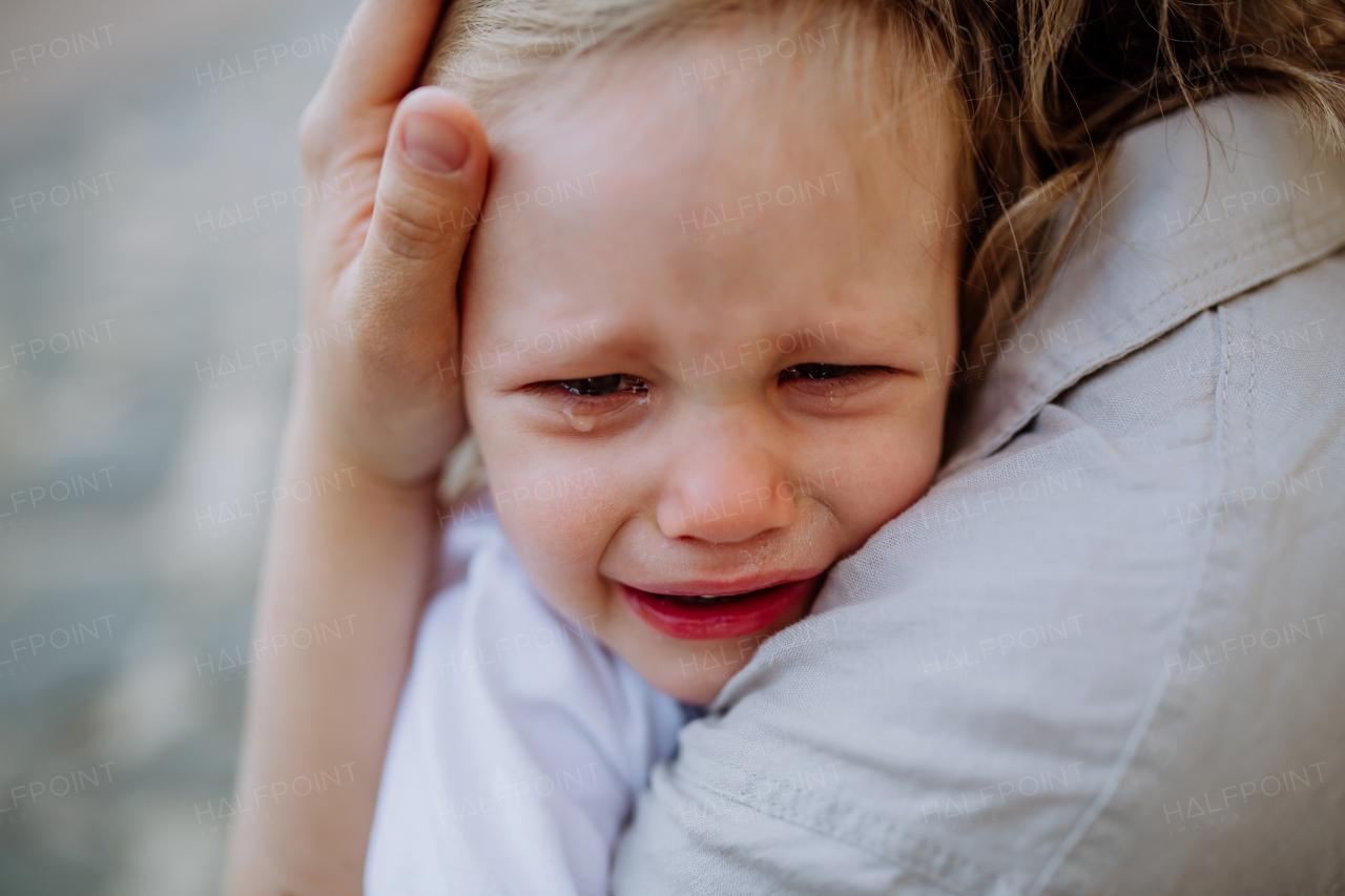 An unrecognizable mother consling her little daughter crying, holding her in arms in street in summer, close-up