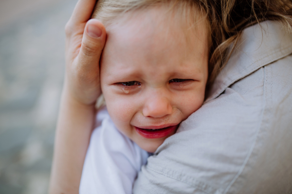 An unrecognizable mother consling her little daughter crying, holding her in arms in street in summer, close-up