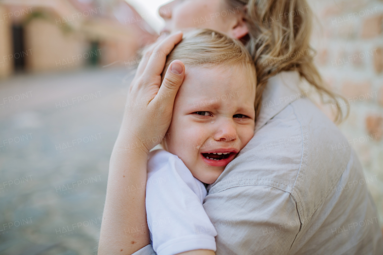 An unrecognizable mother consling her little daughter crying, holding her in arms in street in summer, close-up