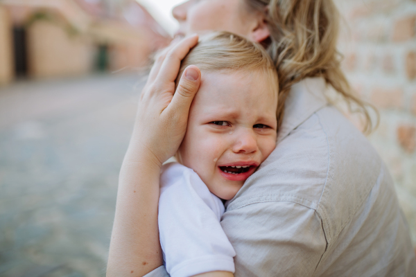 An unrecognizable mother consling her little daughter crying, holding her in arms in street in summer, close-up