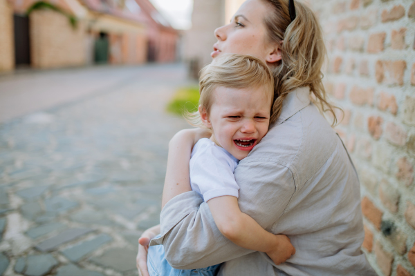 A mother consling her little daughter crying, holding her in arms in street in summer.