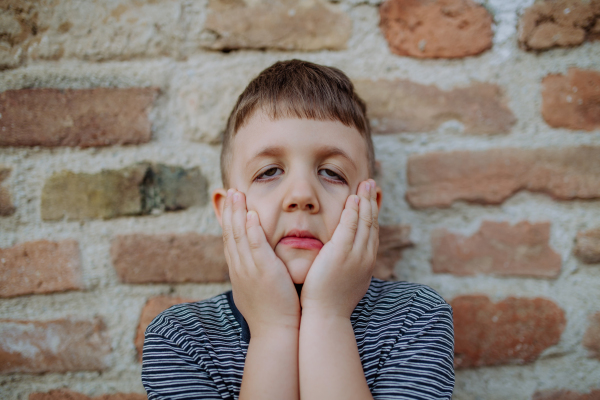 A little boy standing by brick wall and making funny faces in street summer.