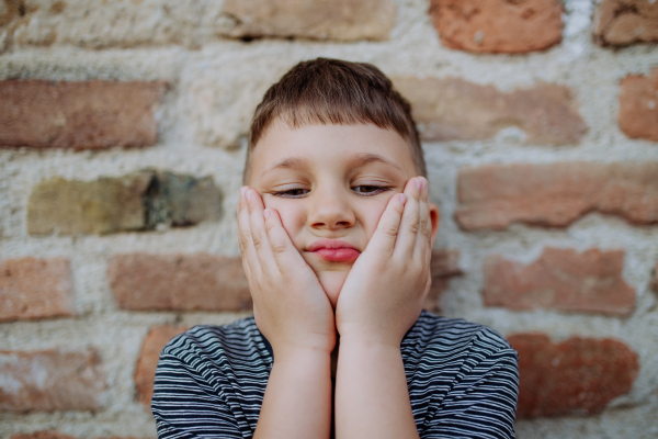 A little boy standing by brick wall and making funny faces in street summer.