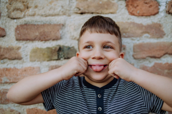 A little boy standing by brick wall and making funny faces in street summer.