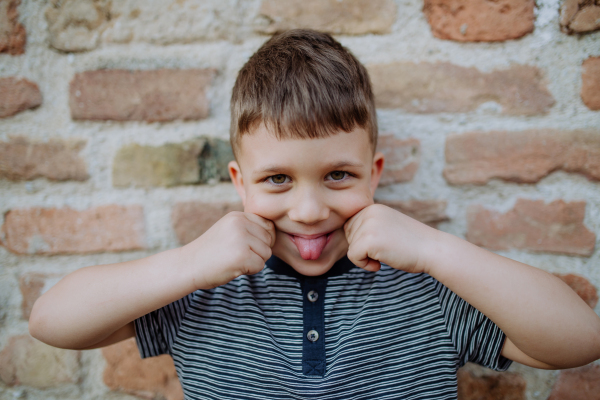 A little boy standing by brick wall and making funny faces in street summer.