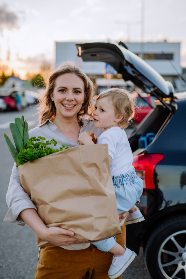 A young mother with little daughter after shopping holding zero waste shopping bags with grocery near car.