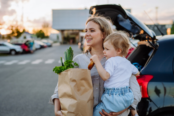 A young mother with little daughter after shopping holding zero waste shopping bags with grocery near car.