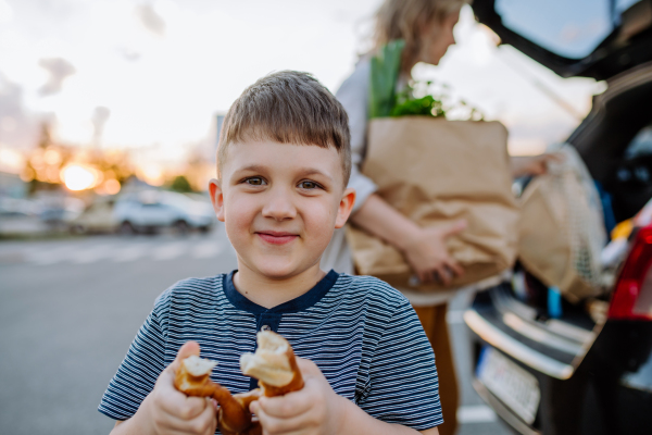 A young mother with little son after shopping holding zero waste shopping bags with grocery, loading car and little boy is eating pastries and looking at camera.