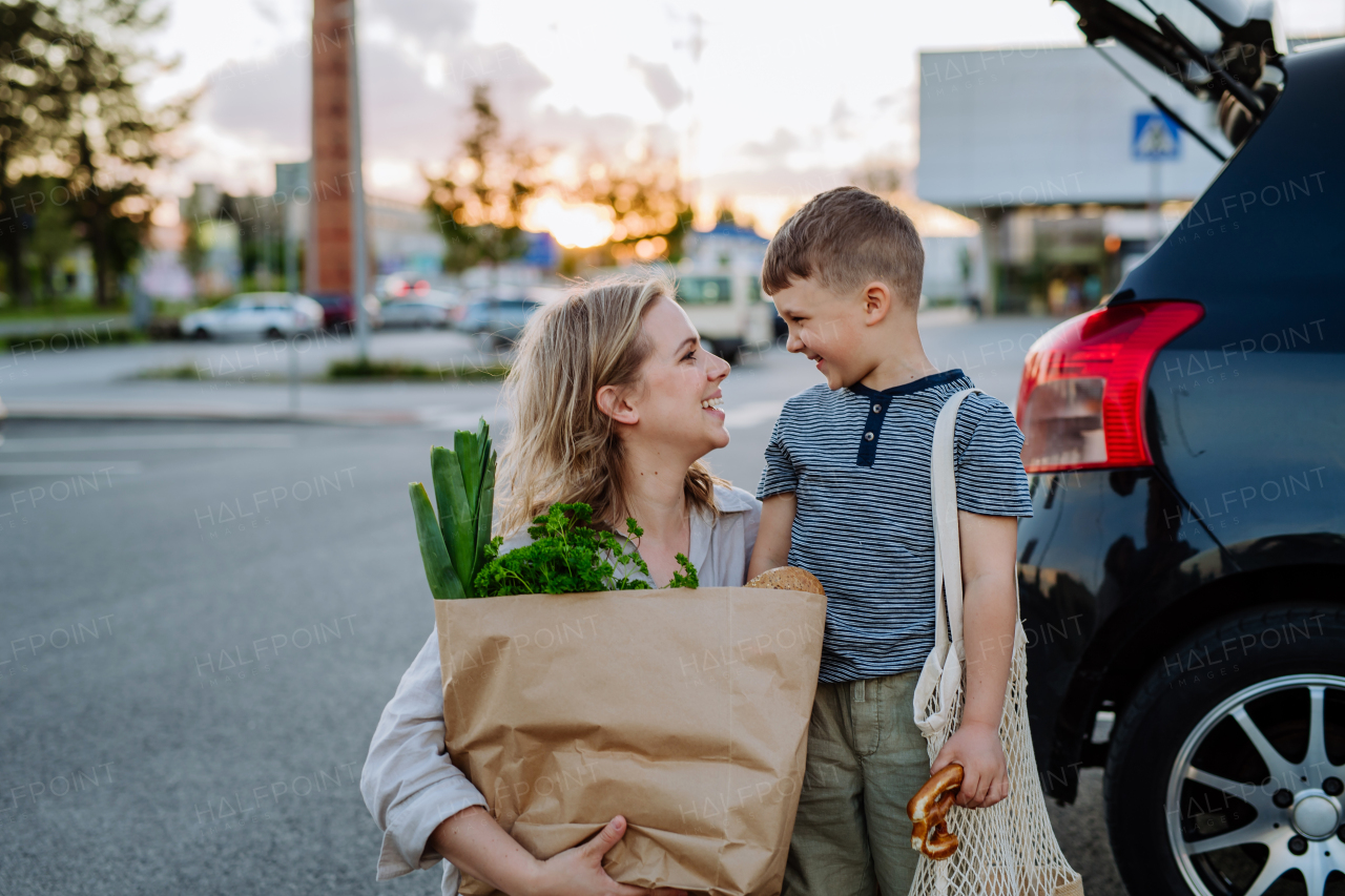 A young mother with little son after shopping holding zero waste shopping bags with grocery near car.