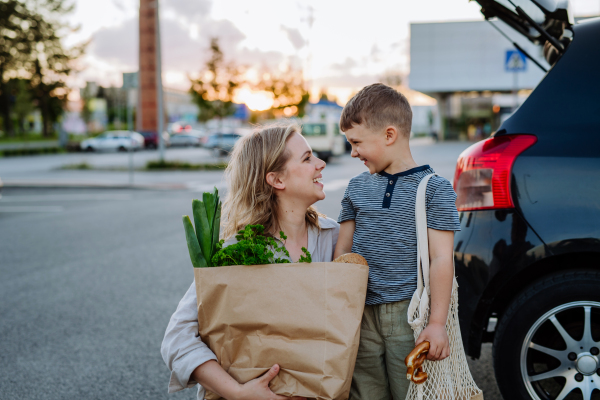 A young mother with little son after shopping holding zero waste shopping bags with grocery near car.