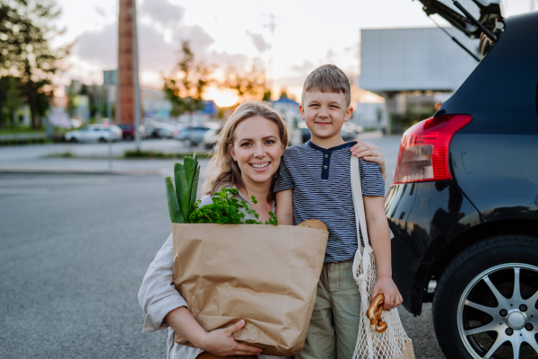 A young mother with little son after shopping holding zero waste shopping bags with grocery near car.