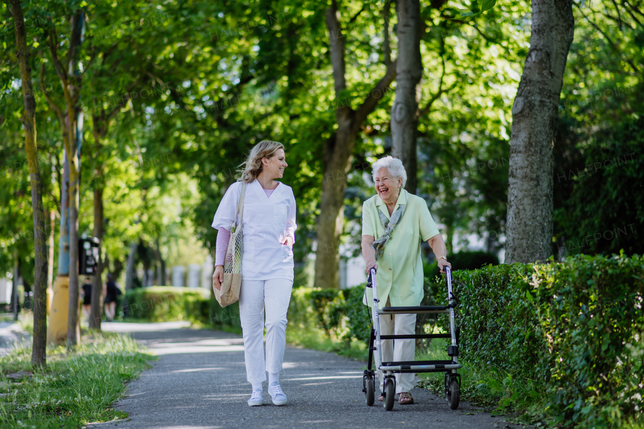 A caregiver with senior woman on walk with walker in park with shopping bag.