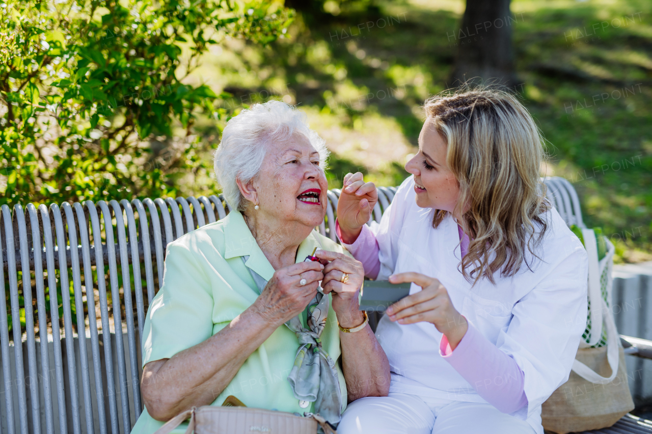 A caregiver helping senior woman to apply lipstick when sitting on bench in park in summer.