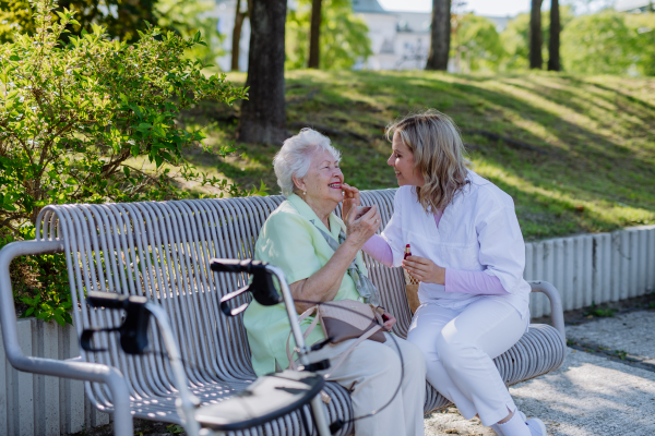 A caregiver helping senior woman to apply lipstick when sitting on bench in park in summer.