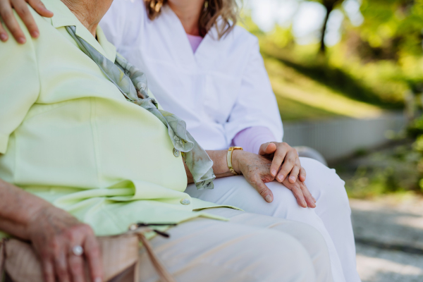 A close-up of caregiver consoling senior woman and touching her hand when sitting on bench in park in summer.