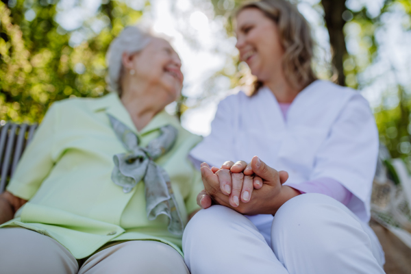 A low angle view of caregiver consoling senior woman and touching her hand when sitting on bench in park in summer.