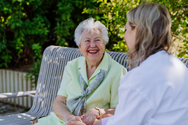 A caregiver with senior woman sitting on bench in park in summer, looking at camera.