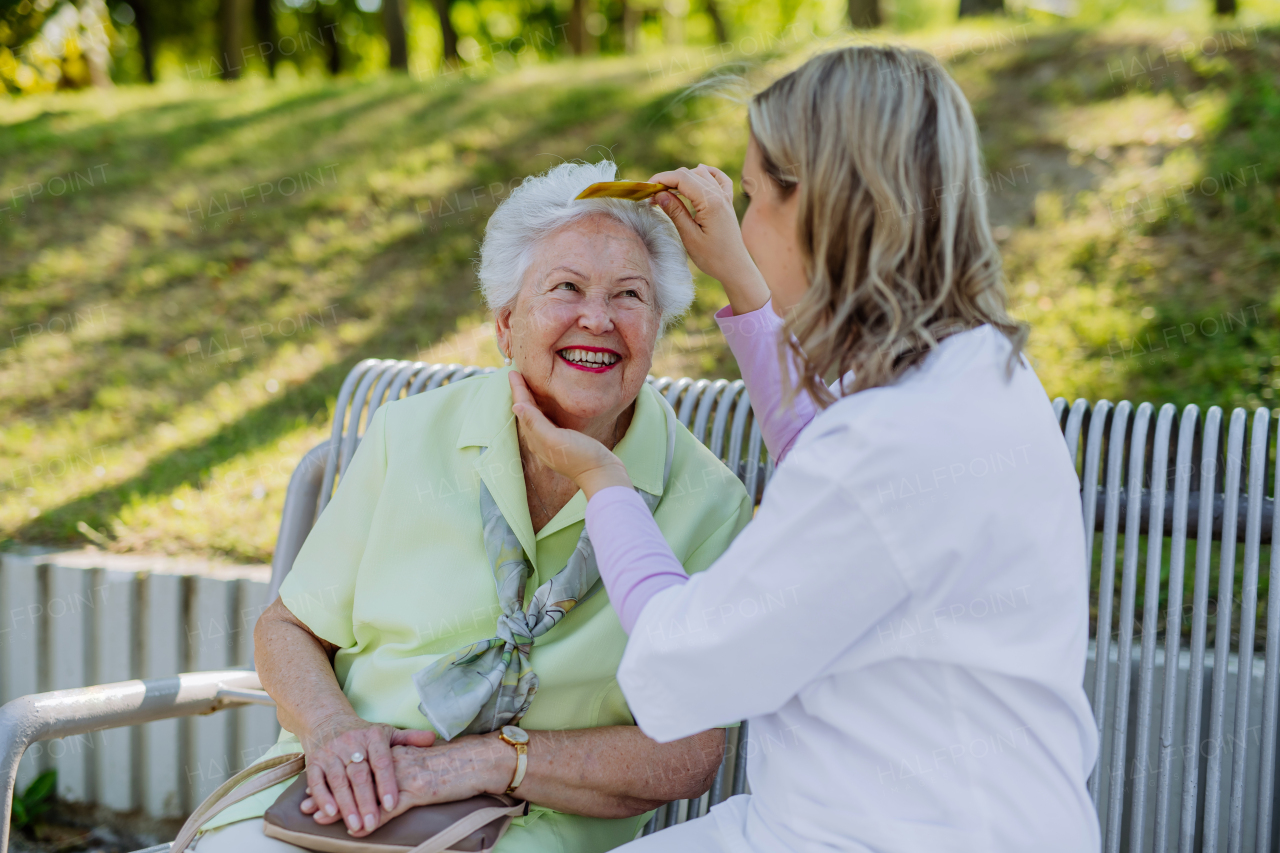 A caregiver helping senior woman to comb hair and make hairstyle when sitting on bench in park in summer.