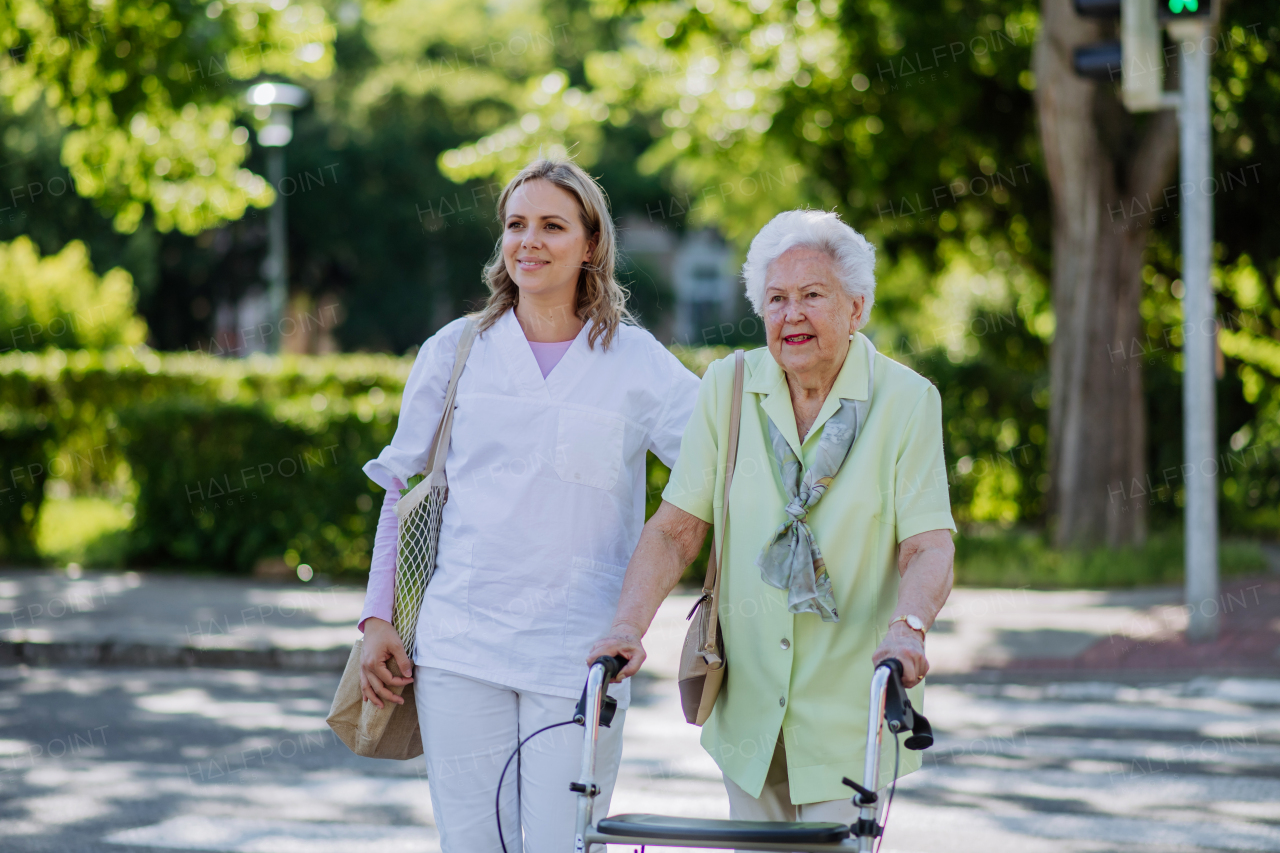 A caregiver with senior woman on walk with walker in park with shopping bag.