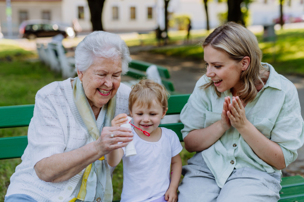 A happy smiling woman with senior grandmother and little daughter blowing soap bubbles at park, family, generation and people concept.