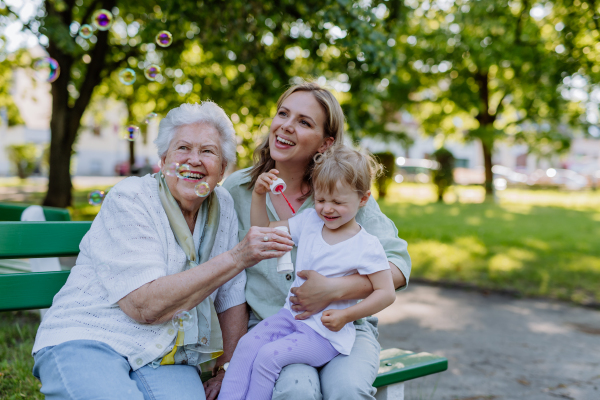 A happy smiling woman with senior grandmother and little daughter blowing soap bubbles at park, family, generation and people concept.