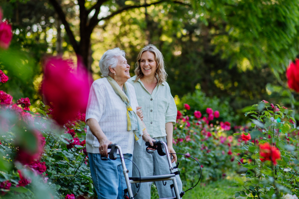 An adult granddaguhter supporting her senior grandmother when taking her for walk with walker in park in summer.