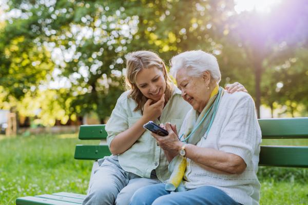 An adult granddaguhter helping her grandmother to use cellphone when sitting on bench in park in summer.