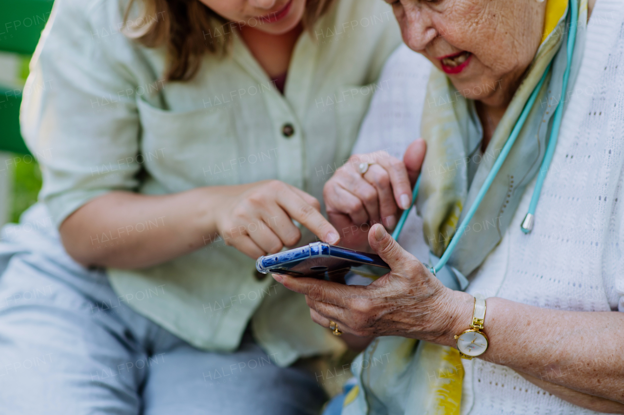 An adult granddaguhter helping her grandmother to use cellphone when sitting on bench in park in summer.