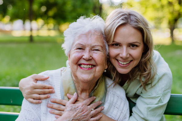 A portrait of adult granddaughter hugging her senior grandmother when sitting on bench in park.