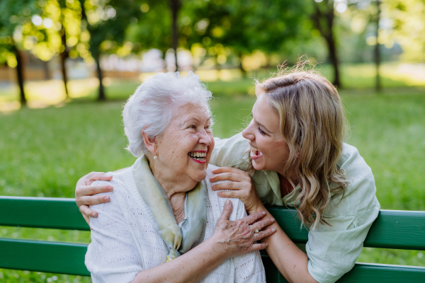 An adult granddaguhter helping her grandmother to use cellphone when sitting on bench in park in summer.