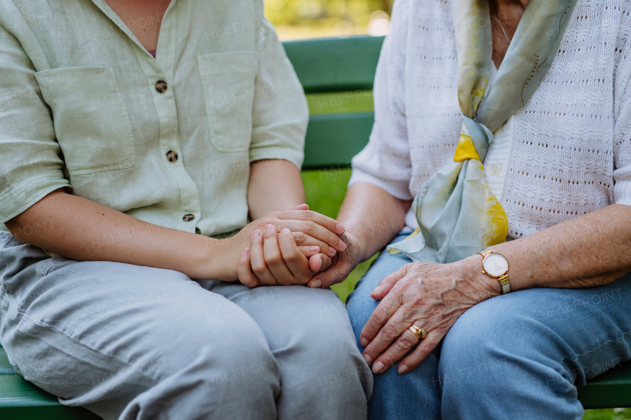 A close-up of granddaughter consoling her senior grandmother and touching her hand when sitting on bench in park in summer.