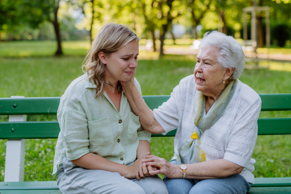 A worried senior grandmother comforting grown up granddaughter when sitting on bench in park, share problem with someone close concept