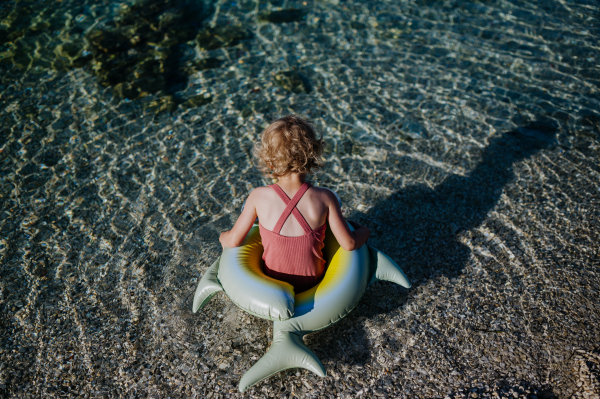 Top view of small girl in pink swimsuit at beach with inflatable toy ring, looking at sea.