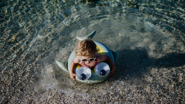 Top view of small girl in pink swimsuit at beach with inflatable toy ring, playing in water looking at sea.