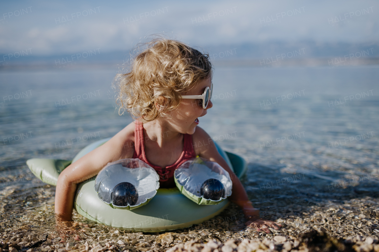 Top view of small girl in pink swimsuit at beach with inflatable toy ring, playing in water looking at sea.