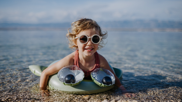 Portrait of small girl in pink swimsuit at beach with inflatable toy ring, playing in water looking at sea.