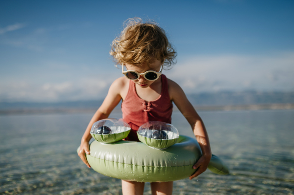 Top view of small girl in pink swimsuit at beach with inflatable toy ring, playing in water looking at sea.