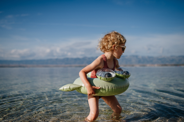 Top view of small girl in pink swimsuit at beach with inflatable toy ring, playing in water looking at sea.