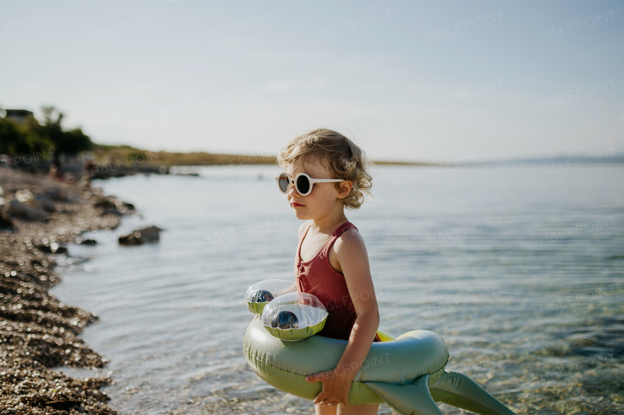 Top view of small girl in pink swimsuit at beach with inflatable toy ring, playing in water looking at sea.