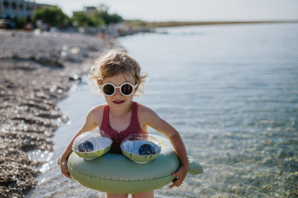 Portrait of small girl in pink swimsuit at beach with inflatable toy ring, playing in water looking at sea.