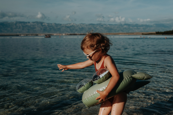 Portrait of small girl in pink swimsuit at beach with inflatable toy ring, playing in water looking at sea.