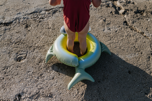 Close up of small girl in red swimsuit standing inflatable toy ring, looking at sea.