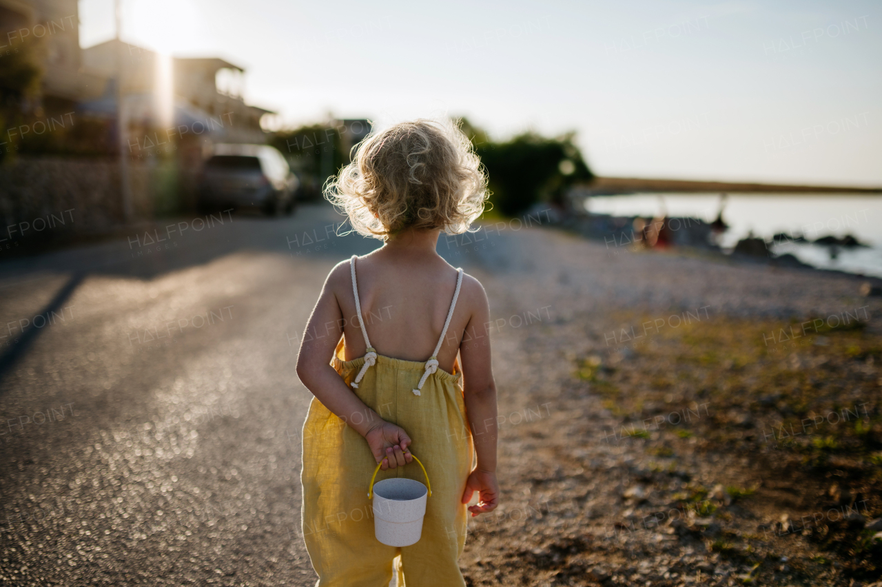 Blonde girl in summer outfit on a walk, holding small plastic bucket, going to search for seashells on beach.