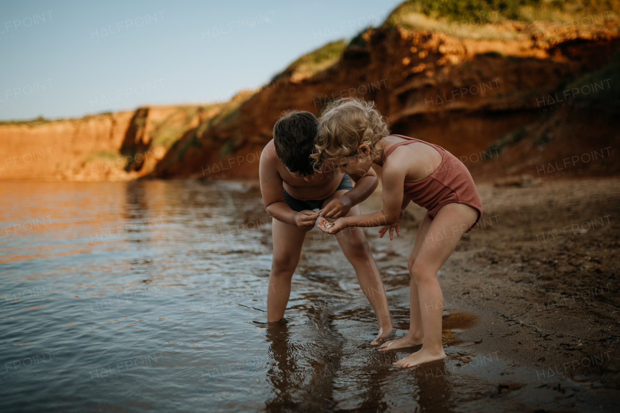 Brother helping sister seraching for shells in wet sand. Small girl in swimsuit playing at beach, crouching in water.