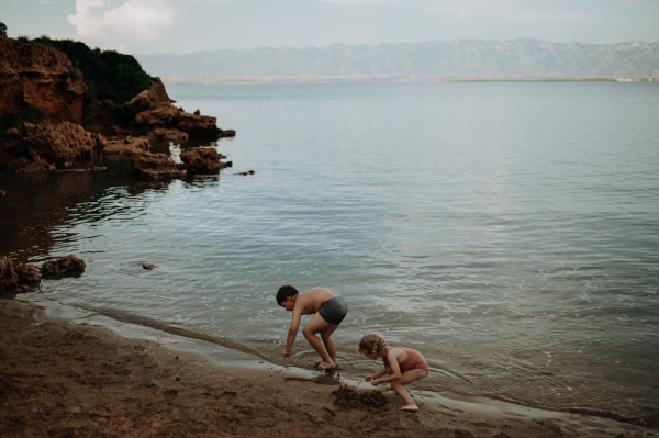 Brother helping sister seraching for shells in wet sand. Small girl in swimsuit playing at beach, crouching in water.