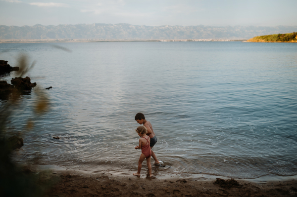 Brother helping sister seraching for shells in wet sand. Small girl in swimsuit playing at beach, crouching in water.