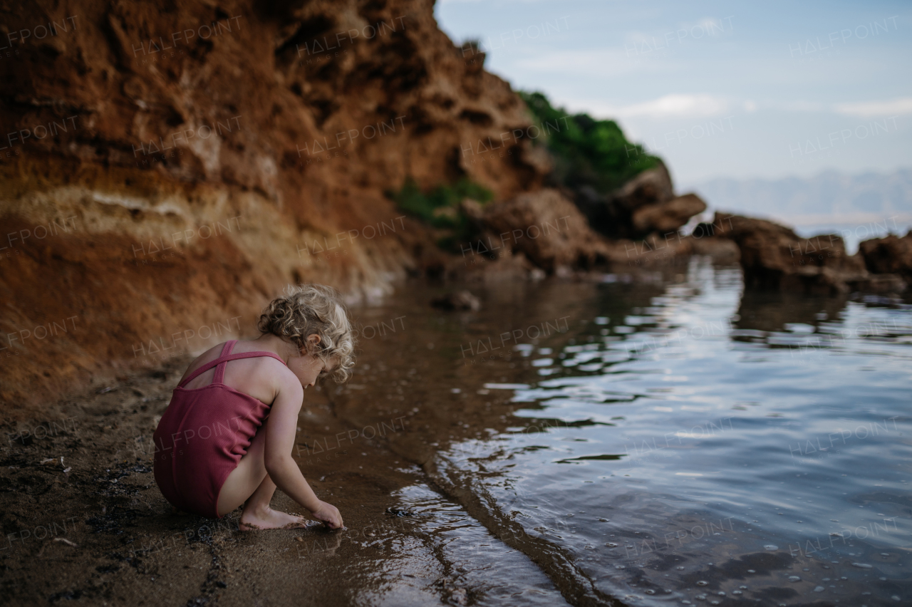 Small girl in swimsuit playing at beach, crouching and seraching for shells in the sand.