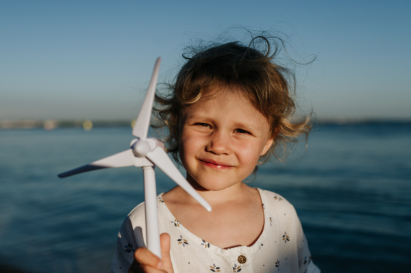 Small girl with blonde hair standing at beach, holding wind turbine model in the hand, sea wind spinning blades.